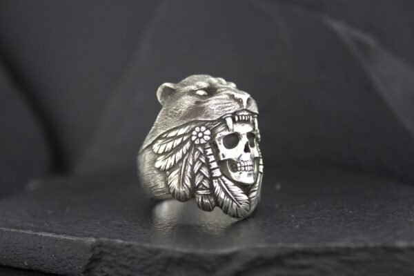 A silver ring featuring a bear and a skull with feathers design.