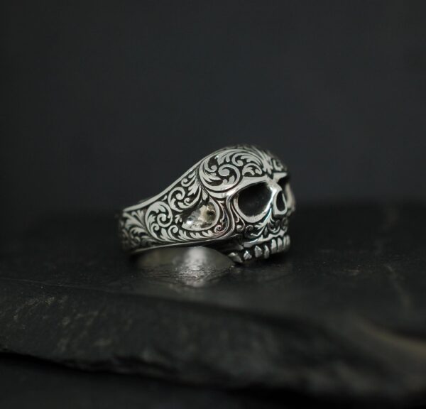 Intricately designed silver skull ring with detailed engravings on a dark background.