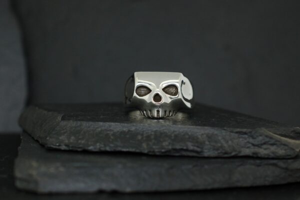 A silver skull ring with a unique design rests on dark textured stones.