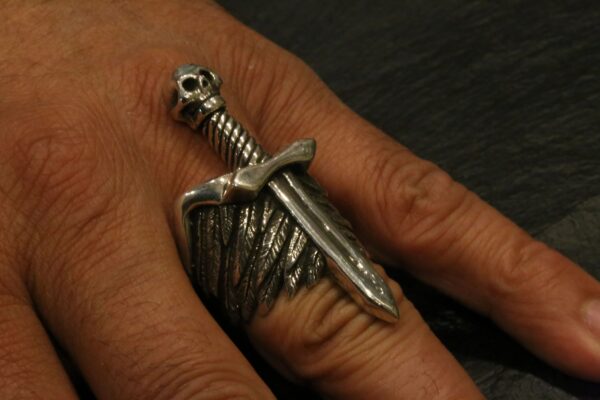 A hand wearing a silver sword-shaped ring with detailed wings.