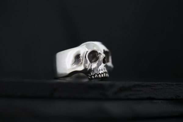 A silver skull ring with detailed eye sockets and teeth on a dark background.