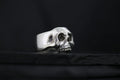 A silver skull ring with detailed eye sockets and teeth on a dark background.