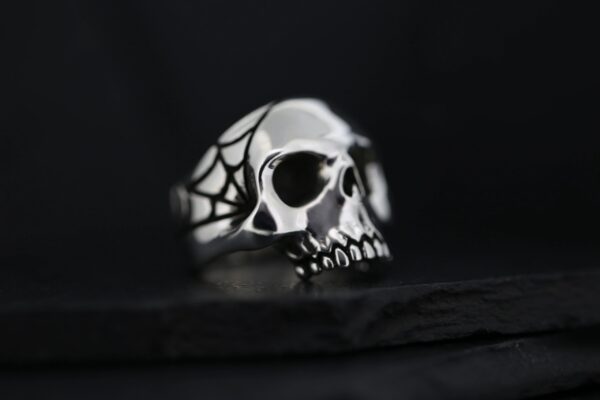 A silver skull ring with spider web details on a dark background.