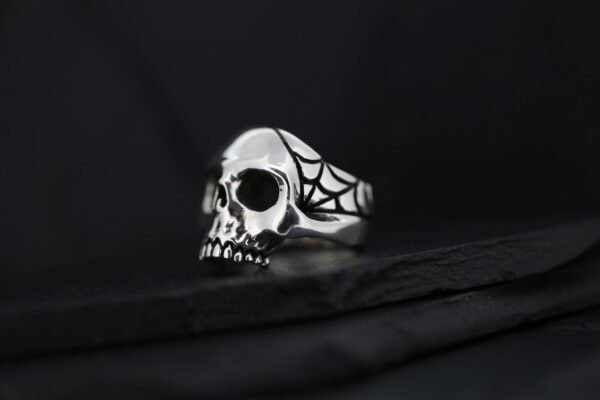 A silver skull ring with a spider web design on a black surface.