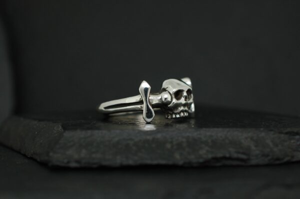 Silver ring featuring a skull and dagger design on a dark background.