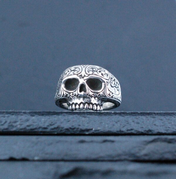 A silver skull ring with intricate details on a dark surface.
