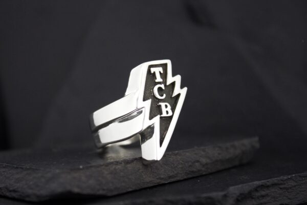 A silver ring with a lightning bolt and 'TCB' initials.