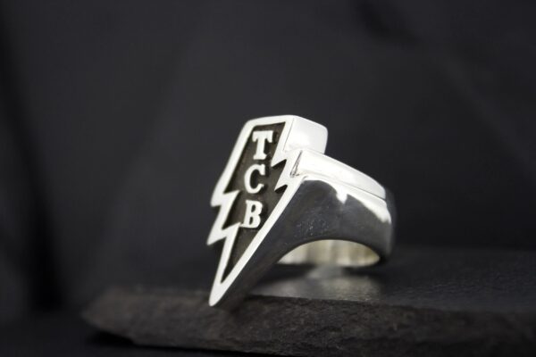 Silver ring with TCB lightning bolt design on a black background.