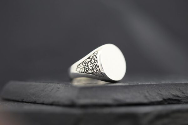 Silver signet ring with intricate side engravings on a dark surface.