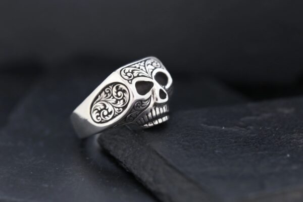 A silver skull ring with intricate side engravings on a dark surface.