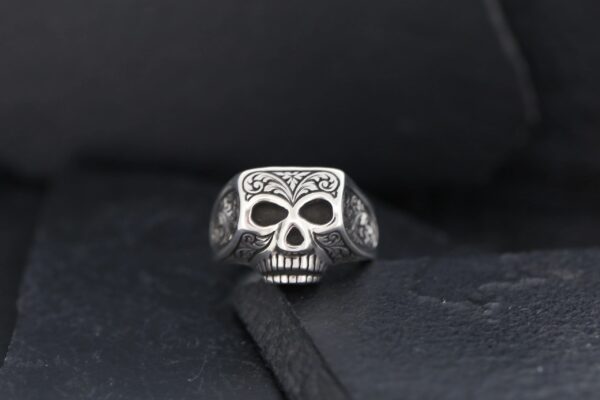 Silver skull ring with intricate designs on a black background.