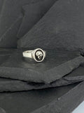 Silver ring with a skull design resting on dark slate stones.