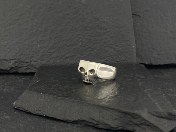 Silver skull-shaped ring with hollow eyes on a black textured surface.