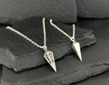 Two silver geometric pendant necklaces on a dark stone surface.