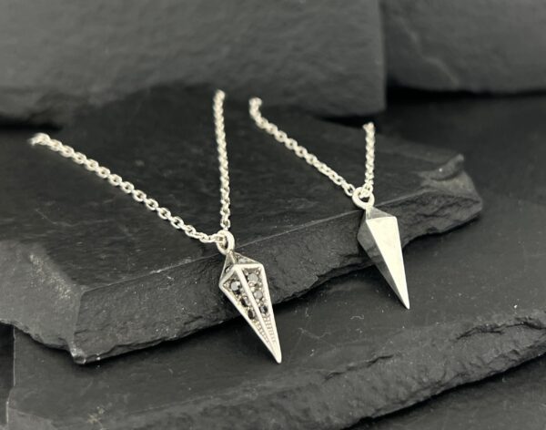 Two silver geometric pendant necklaces on a dark stone surface.