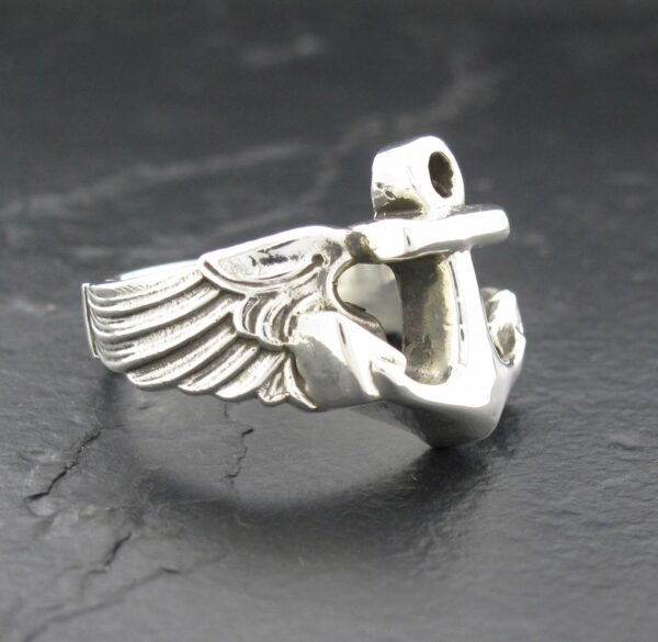 Silver ring with anchor and wing design on a dark surface.