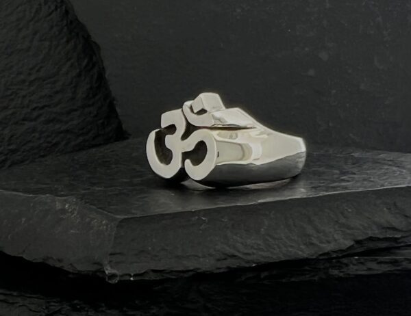 Silver ring featuring an Om symbol design on a black textured surface.