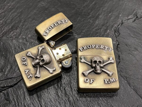 Two skull-themed brass lighters with inscriptions on a black surface.