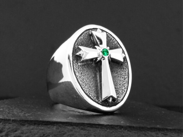 Silver ring with a cross and green gem on a black background.