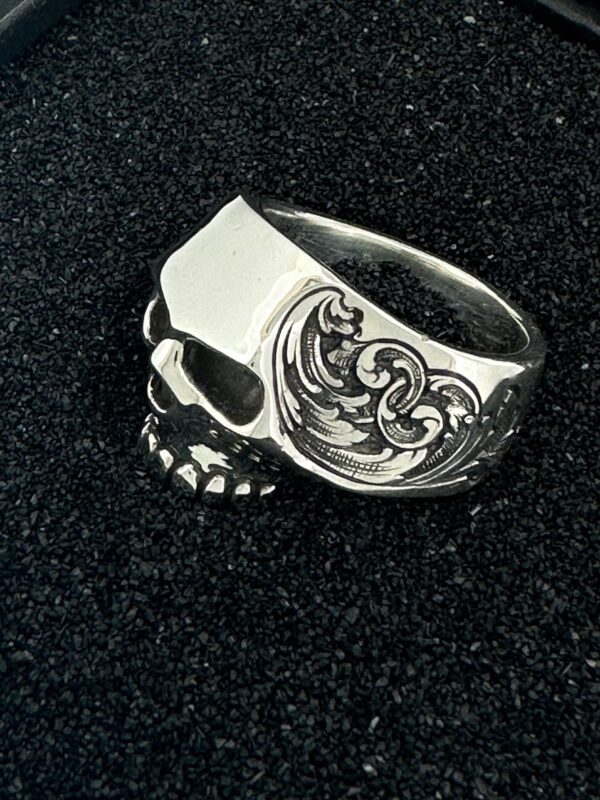 Silver skull ring with intricate side engravings on black background.