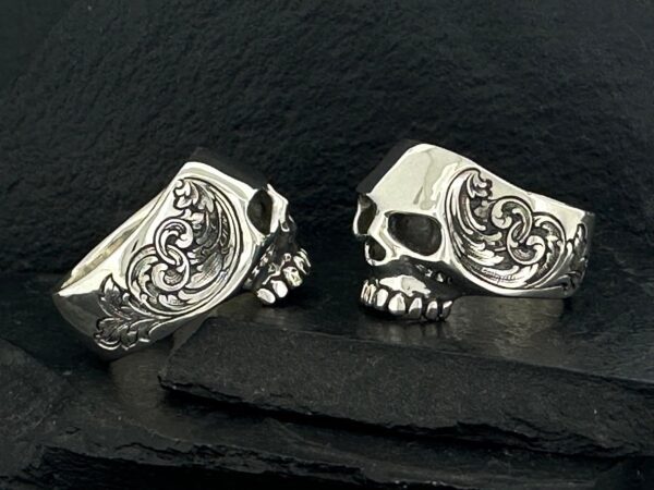 Two intricately designed silver skull rings on a dark surface.