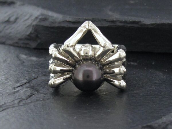 Silver spider ring holding a black pearl on a dark stone background.