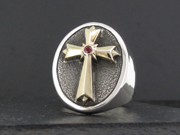 A silver ring featuring a gold cross with a red gemstone at its center.
