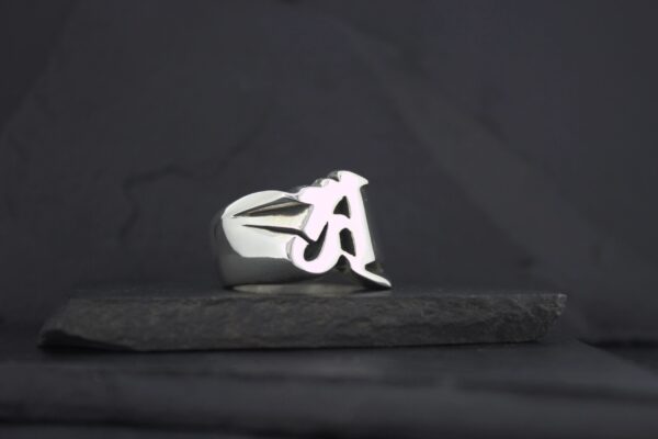 Silver ring with a stylized letter A on a dark background.