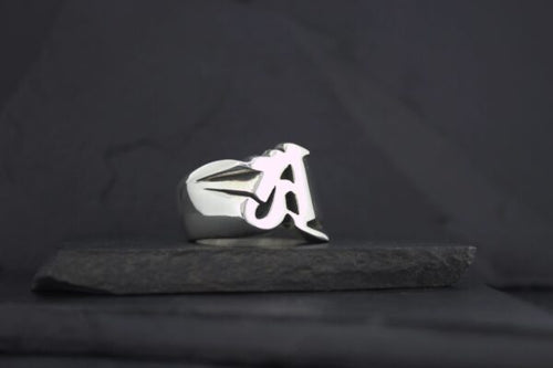 Silver ring with a stylized letter A on a dark background.