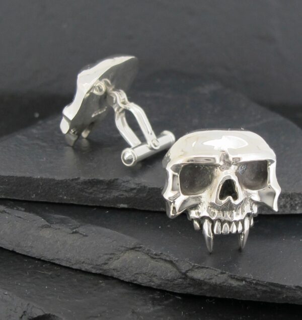 Silver skull cufflinks with detailed fangs on a dark slate surface.