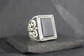 Silver ring with a rectangular black stone and ornate side details.