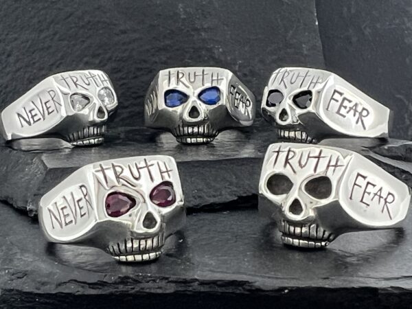 Five silver skull rings with engraved words and colored gems.