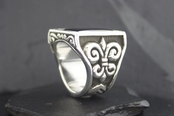 Silver ring with intricate fleur-de-lis design on a black background.