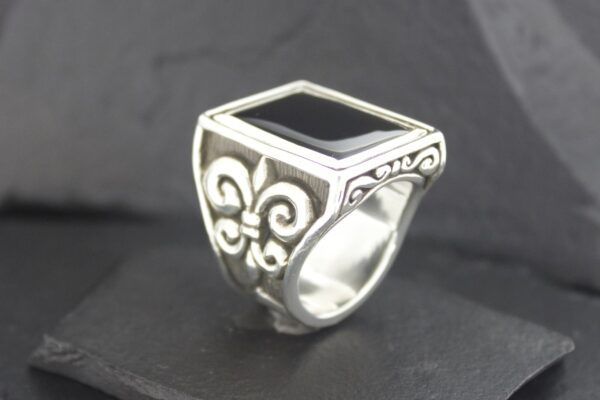 Silver ring with a black rectangular stone and ornate fleur-de-lis designs.
