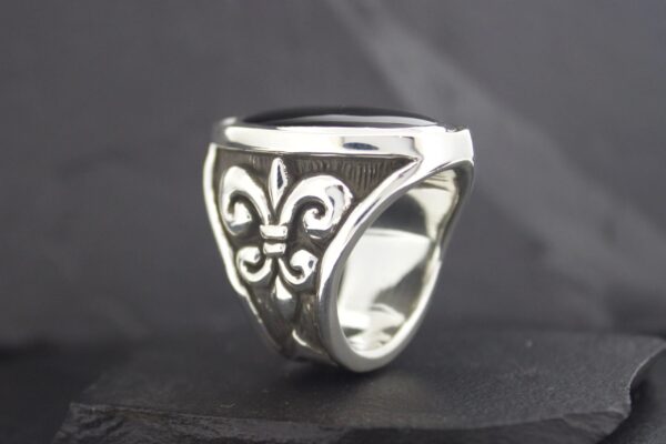 Silver ring with a fleur-de-lis design on a dark background.