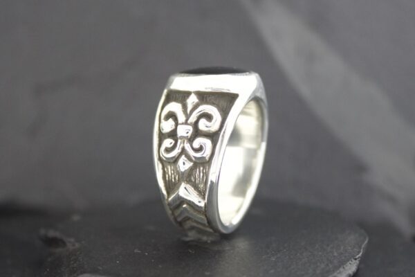 Silver ring with a fleur-de-lis design on a black background.