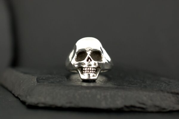 A silver skull ring with detailed facial features on a dark surface.