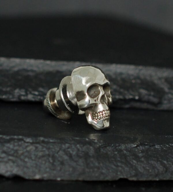 A metallic skull-shaped stud earring on a dark textured surface.