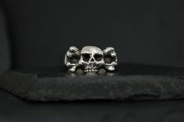 A silver skull ring with crossed bones on a dark background.