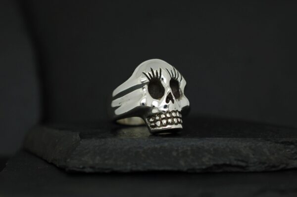 A skull-shaped ring with long eyelashes on a dark surface.