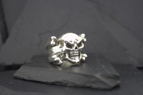 A silver skull ring with crossbones detail on a dark surface.