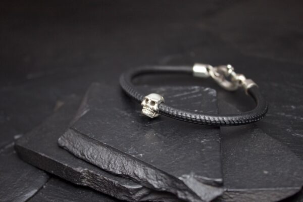 Black leather bracelet with a small silver skull charm on a slate surface.