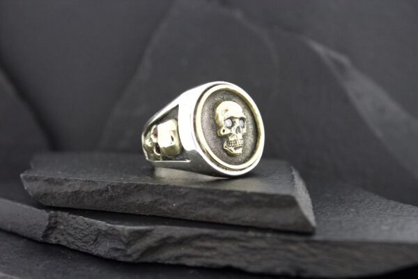 Silver ring with a skull design on a black textured surface.