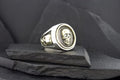 Silver ring with a skull design on a black textured surface.