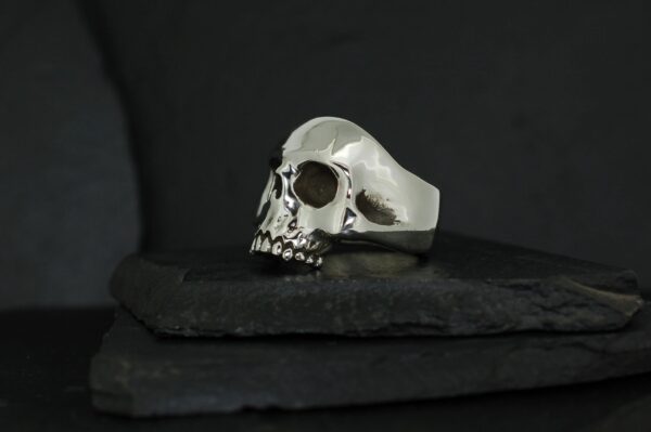 Silver skull-shaped ring displayed on black stone slabs.